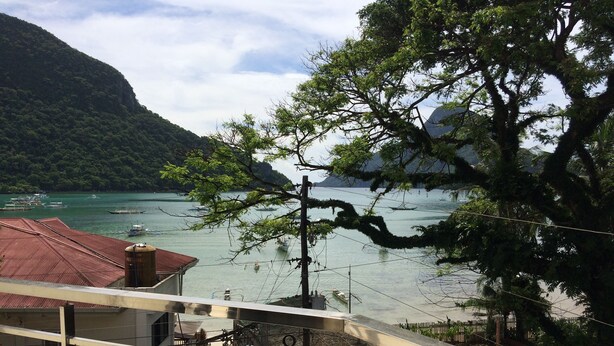 A coastal view with boats on the water, a mountain in the background, and a building with a red roof in the foreground. - Inngo Tourist Inn (El Nido)