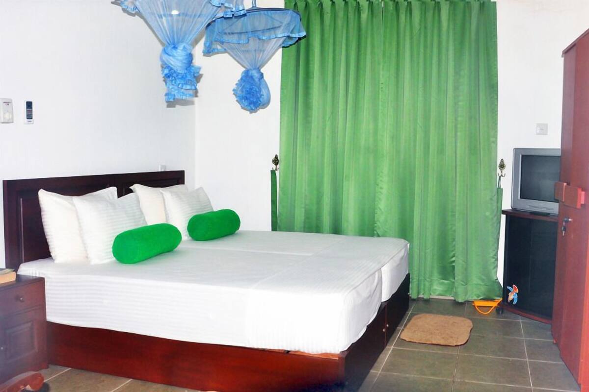 A neatly made bed with white and green pillows, a green curtain, a wooden nightstand, a television on a cabinet, and a brown mat on the floor.