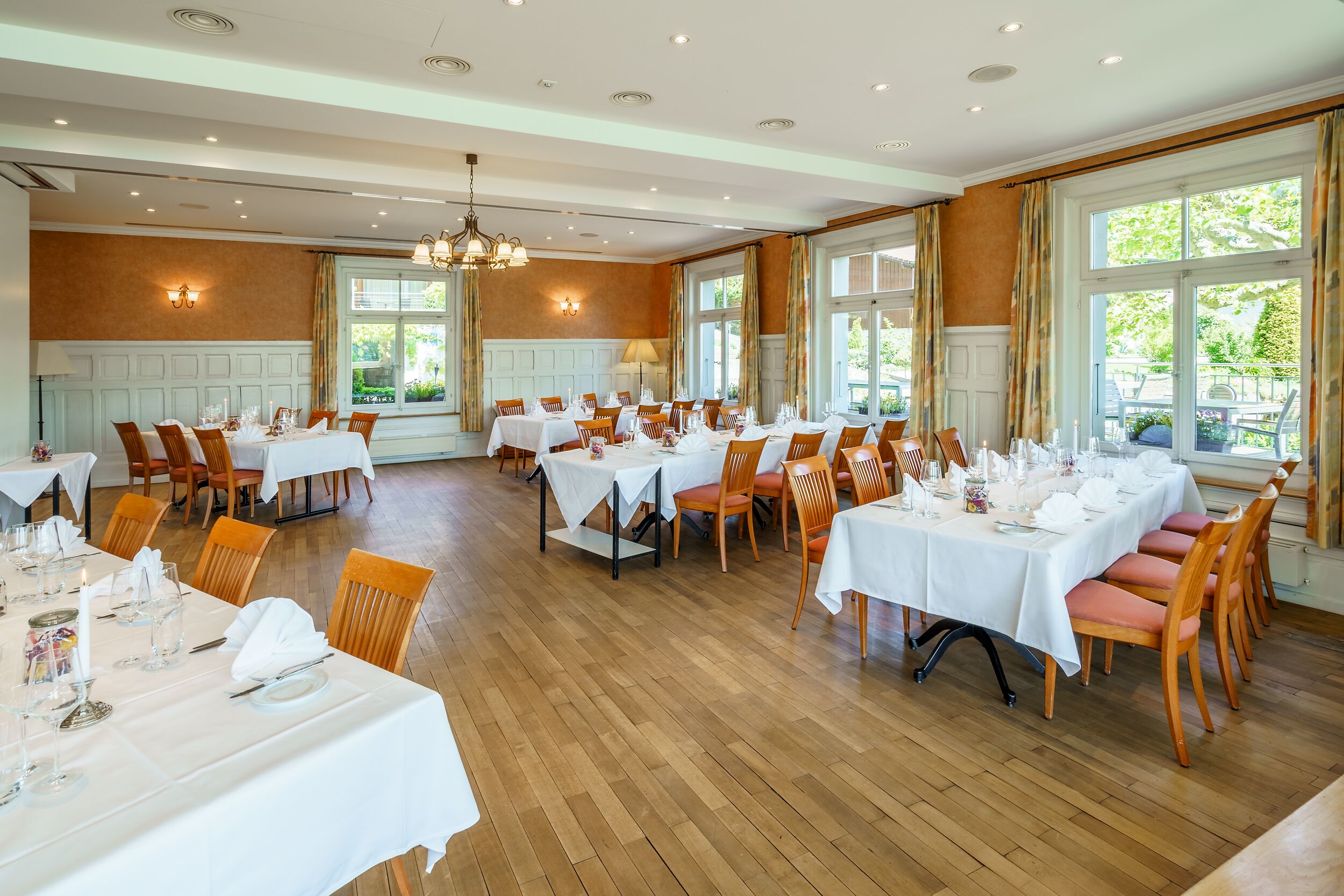 A spacious dining area with multiple tables set for a meal, large windows with curtains, and a wooden floor.