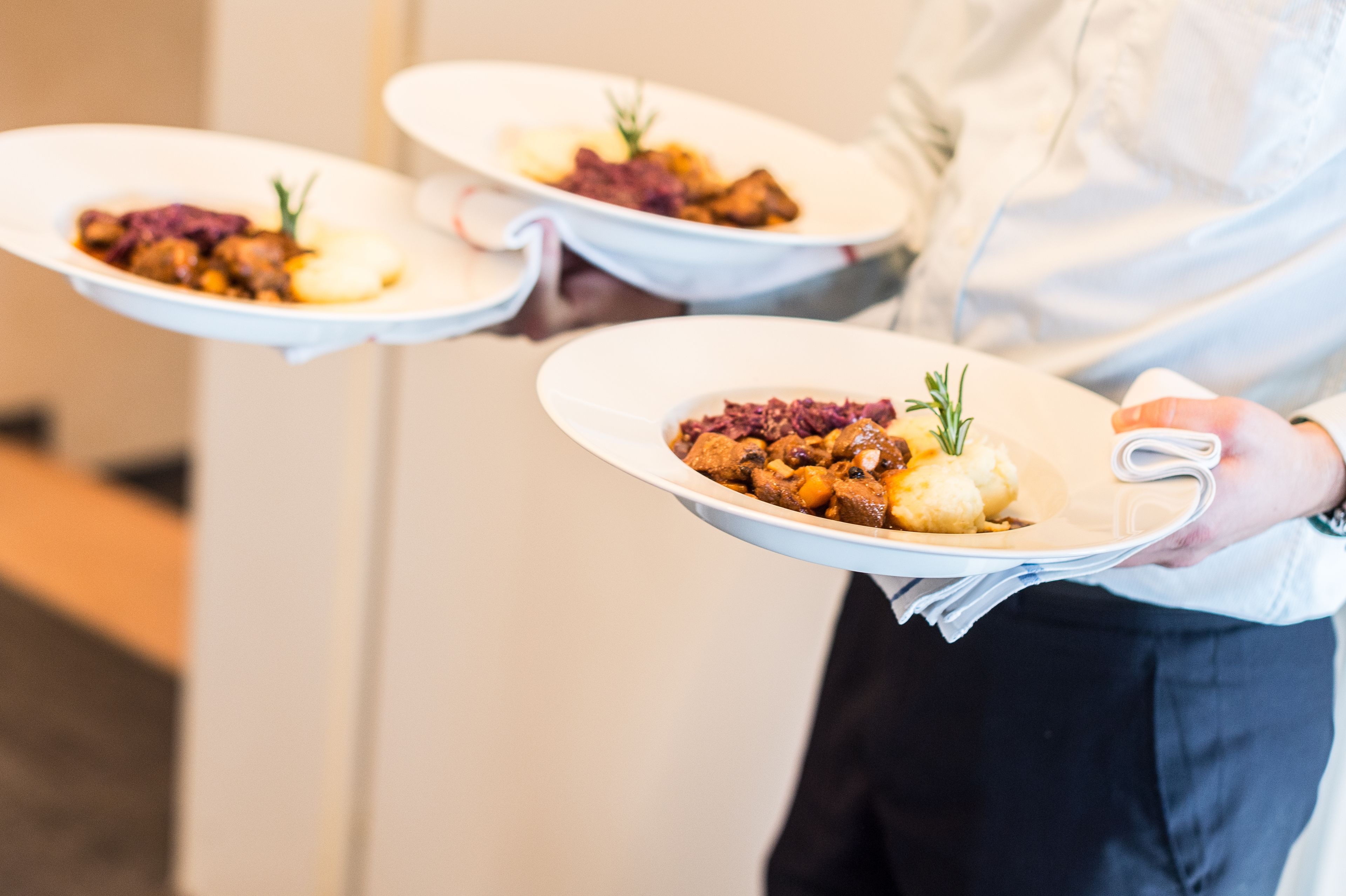 Eine Person hält drei weiße Teller mit Essen, darunter ein Gericht mit Fleisch, Kartoffelpüree und Gemüse.