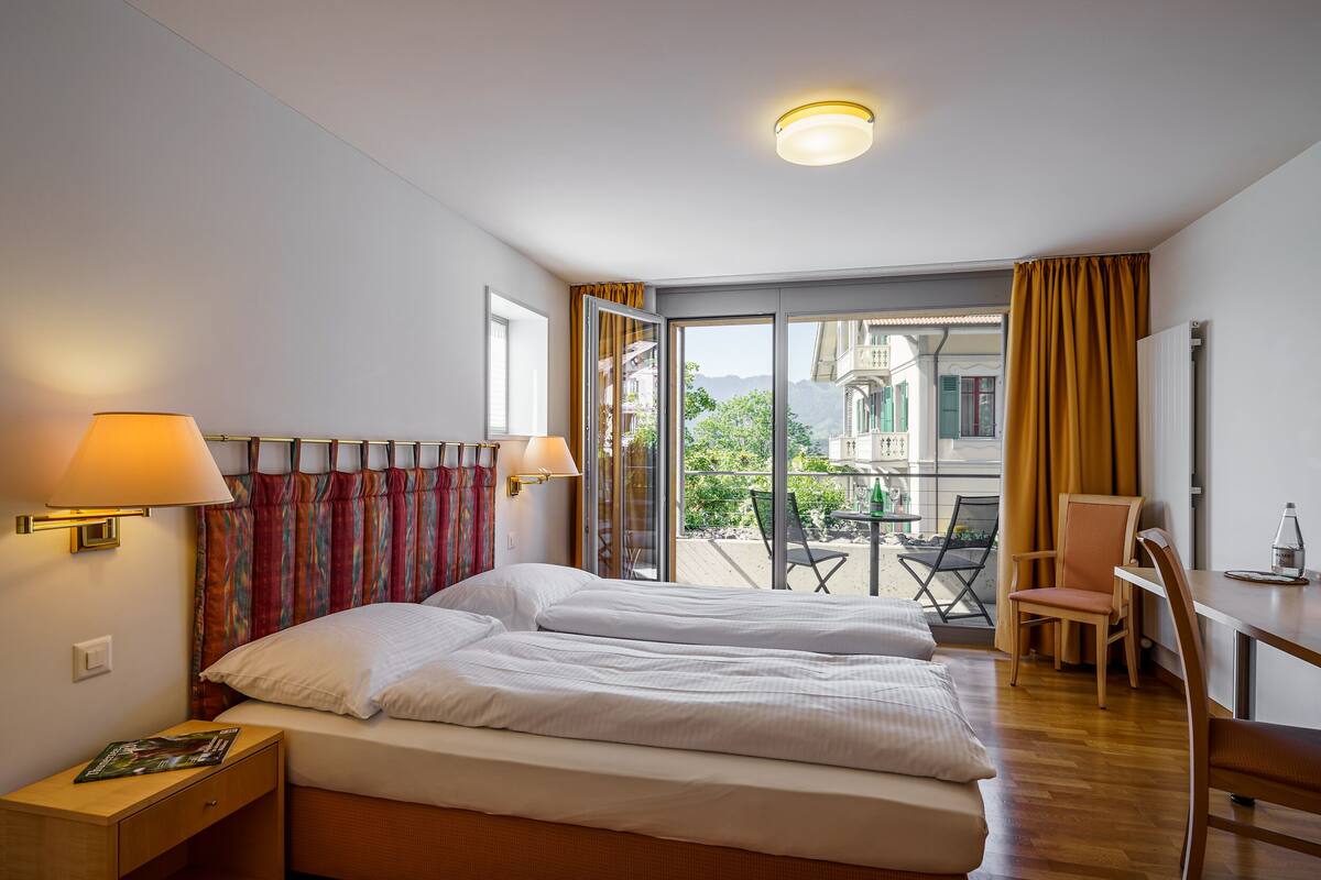 Een hotelkamer met een bed, een bureau en een stoel. Er is een raam met gordijnen en uitzicht op buiten.
