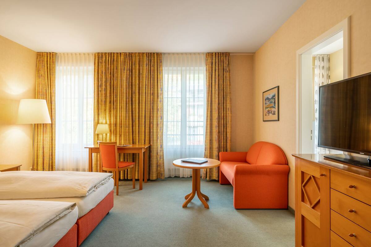 Een hotelkamer met een bed, een bureau met stoel, een oranje fauteuil, een klein rond tafeltje en een televisie op een houten kast.