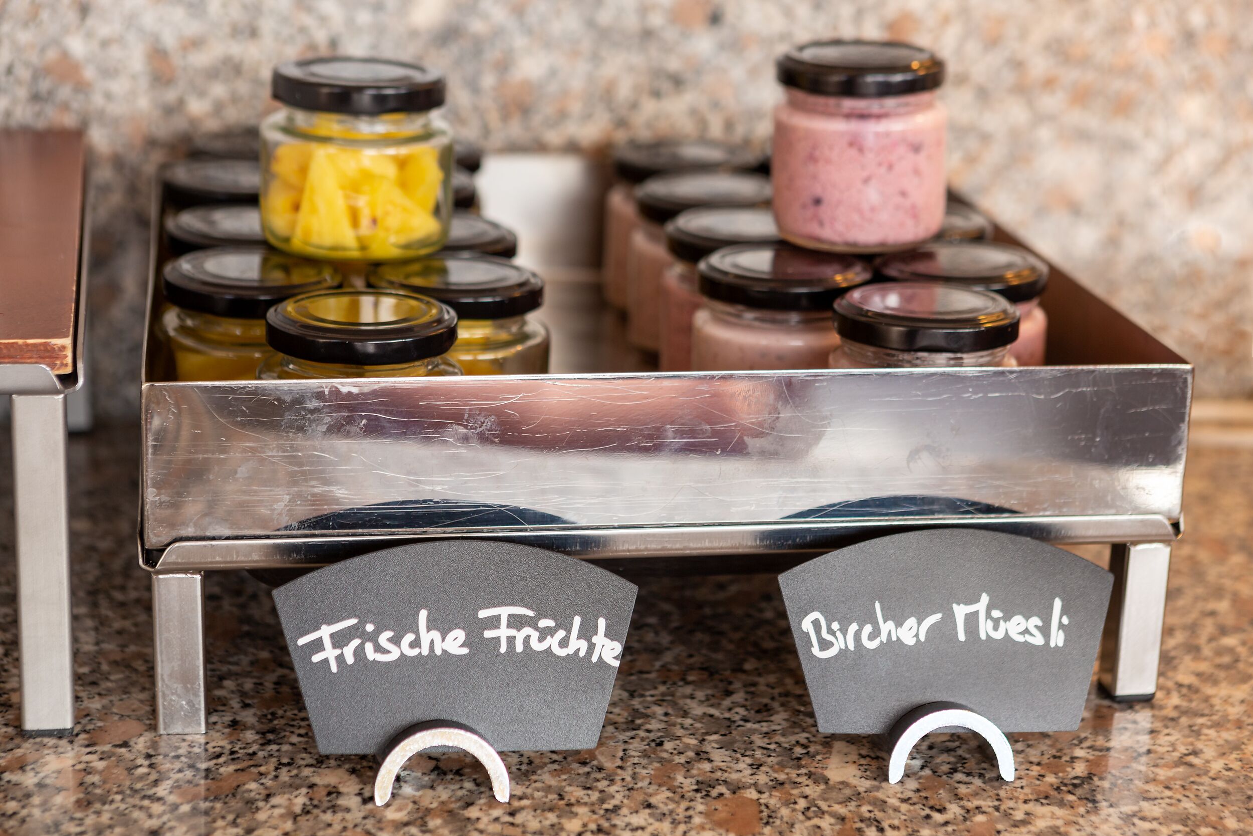 A stainless steel cart with jars of preserves labeled "Frische Fruchte" and "Bacher Muesli" on a granite countertop.