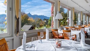 Een restaurant met gedekte tafels, grote ramen met gordijnen en uitzicht op bergen en bomen.