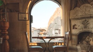 A cozy room with a stone fireplace, a wooden table with two chairs, and a large arched window overlooking a cityscape.