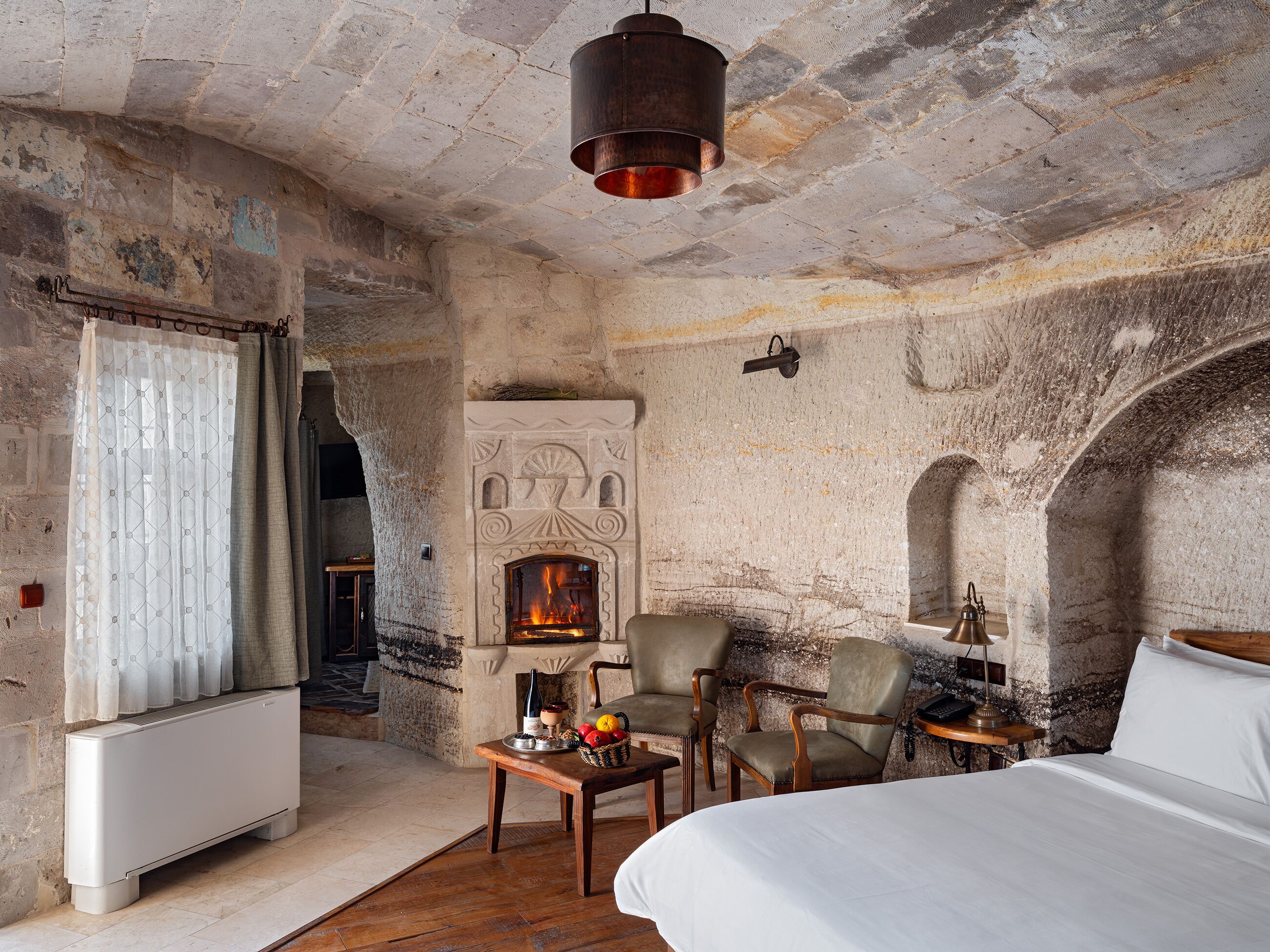 A rustic room with a stone fireplace, a bed, a table with fruit, and two chairs.