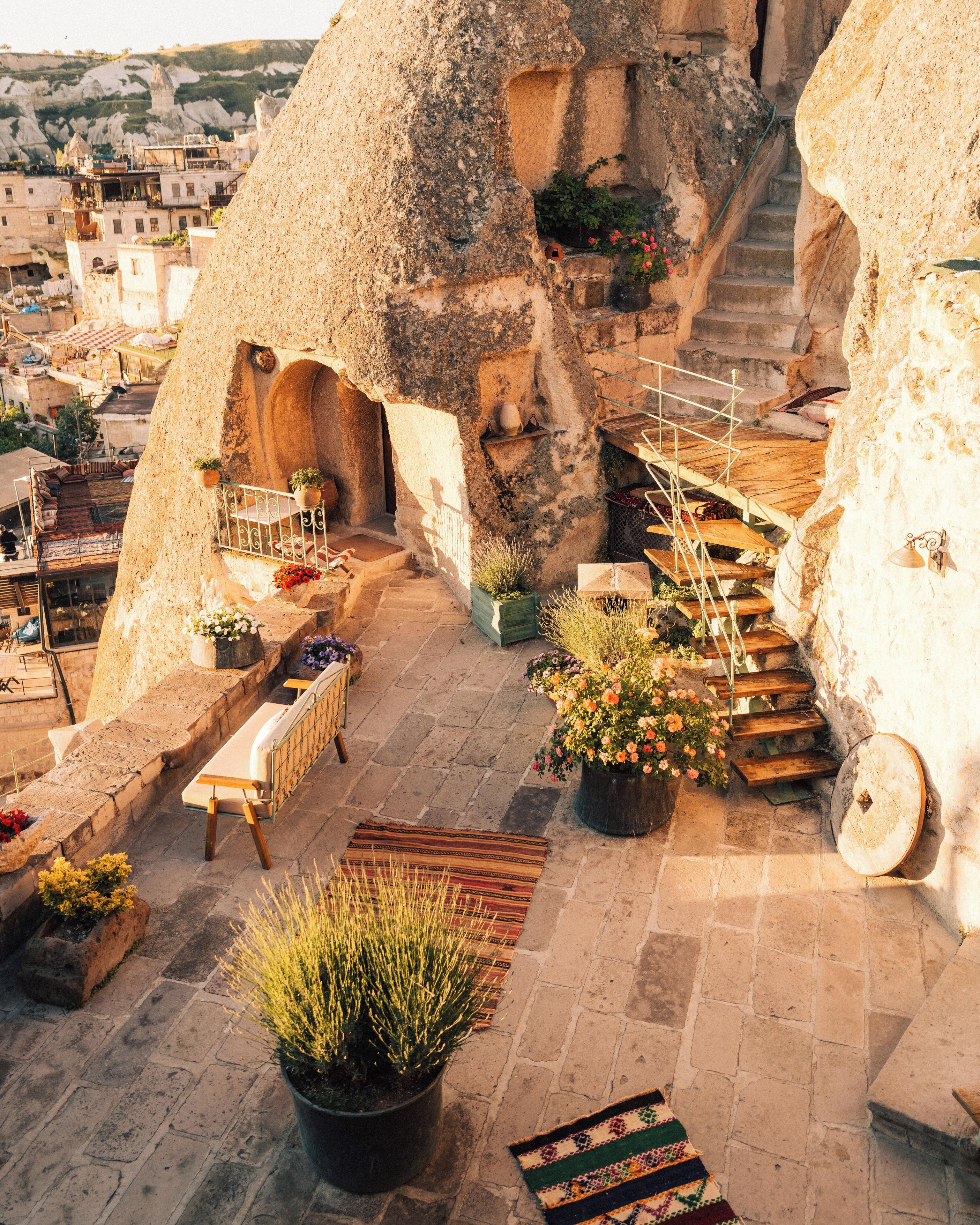 A rooftop terrace with stone walls, a wooden bench, potted plants, and a view of the city.
