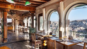 Un comedor rústico con mesas y sillas de madera, paredes de piedra y ventanas arqueadas con vistas a un paisaje urbano.