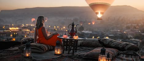 A person sitting on a bed with a table and lanterns, overlooking a cityscape with hot air balloons in the background.