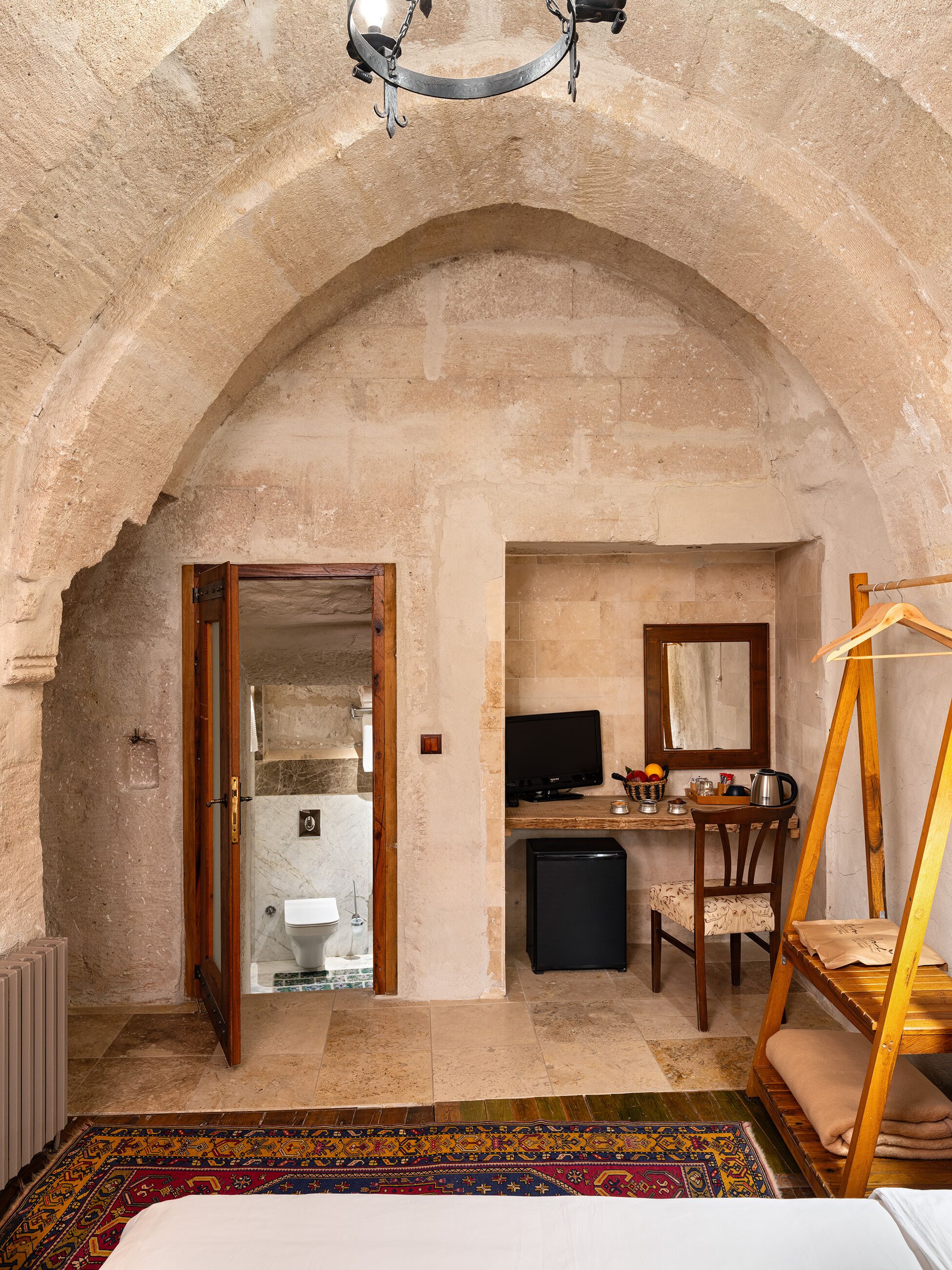 A room with an arched stone ceiling, a bed with a colorful rug, a desk with a computer, a chair, and a wooden swing.
