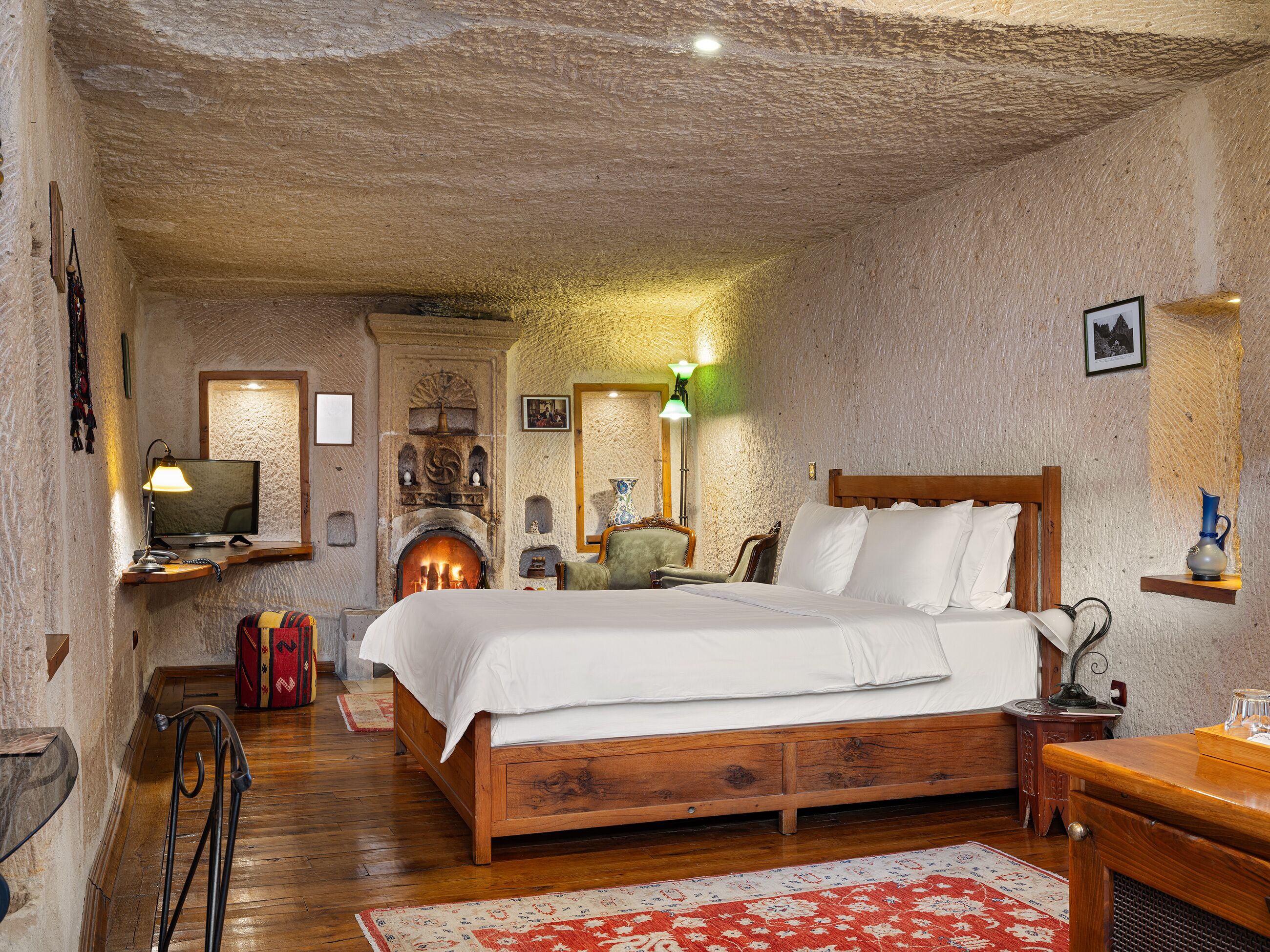 A rustic bedroom with a wooden bed, a fireplace, a television, and a small table with a lamp.
