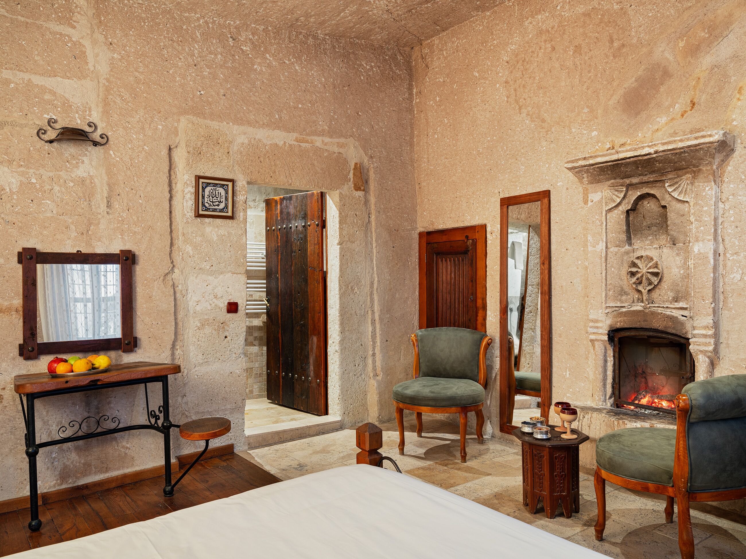 A rustic room with a bed, a wooden table with fruits, a green armchair, a fireplace, and a mirror.
