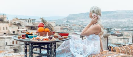 A woman in a white dress sitting on a patterned cushion with a table of food and drinks in front of her, overlooking a cityscape.