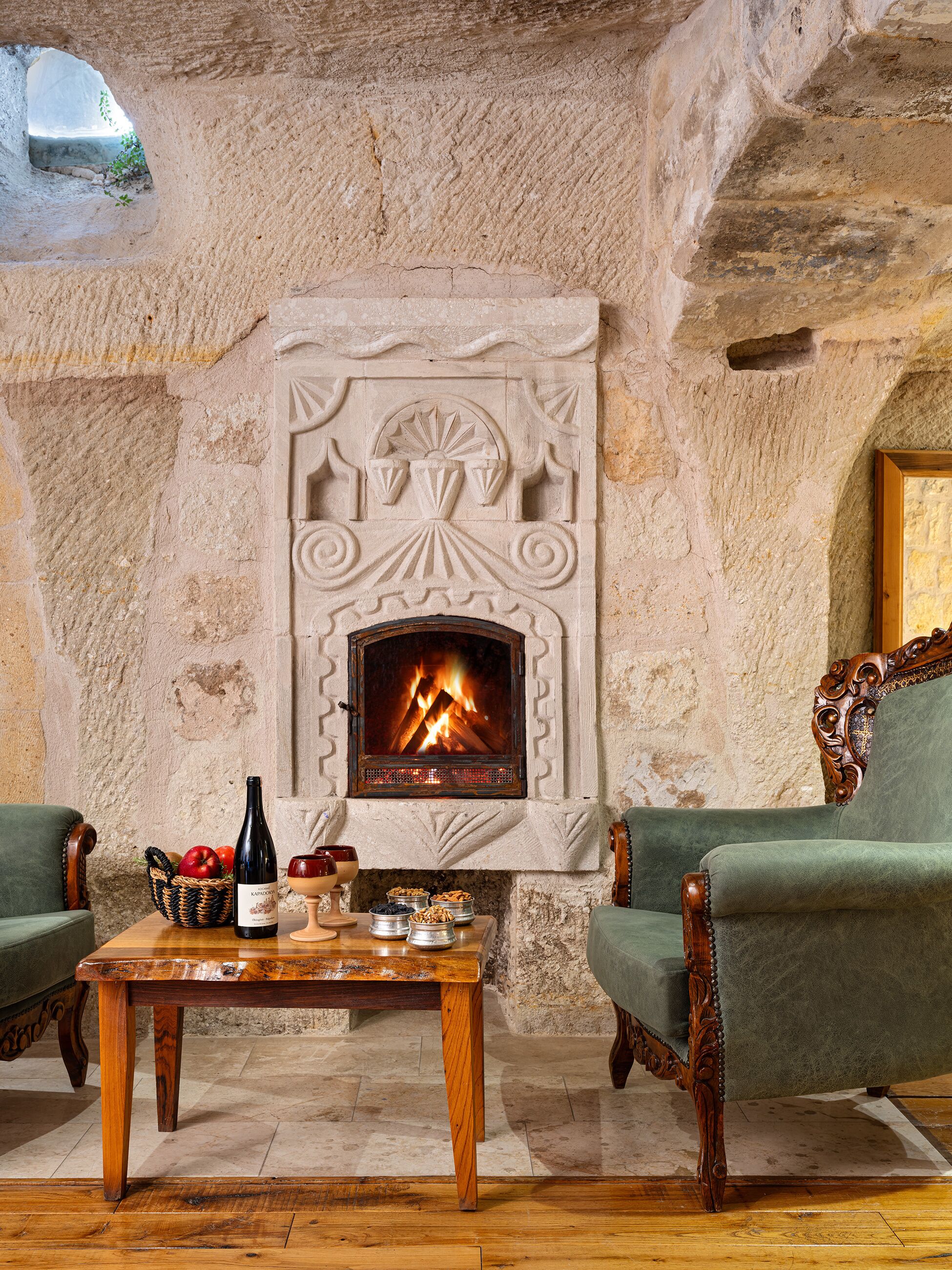 Une cheminée en pierre avec un feu, flanquée de deux fauteuils à accoudoirs recouverts de tissu vert et d’une table en bois sur laquelle se trouvent une bouteille et des en-cas.