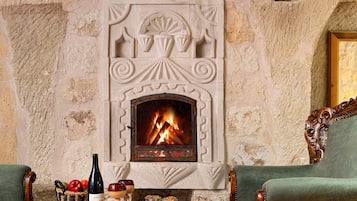Une cheminée en pierre avec un feu, flanquée de deux fauteuils à accoudoirs recouverts de tissu vert et d’une table en bois sur laquelle se trouvent une bouteille et des en-cas.