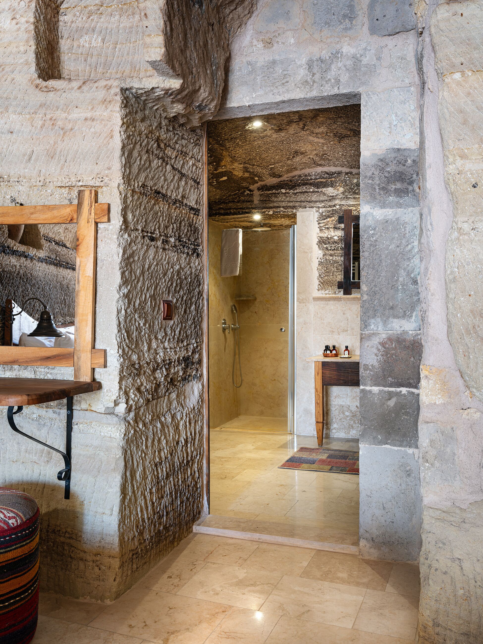 Baño de paredes de piedra con ducha, repisa de madera y alfombra estampada.