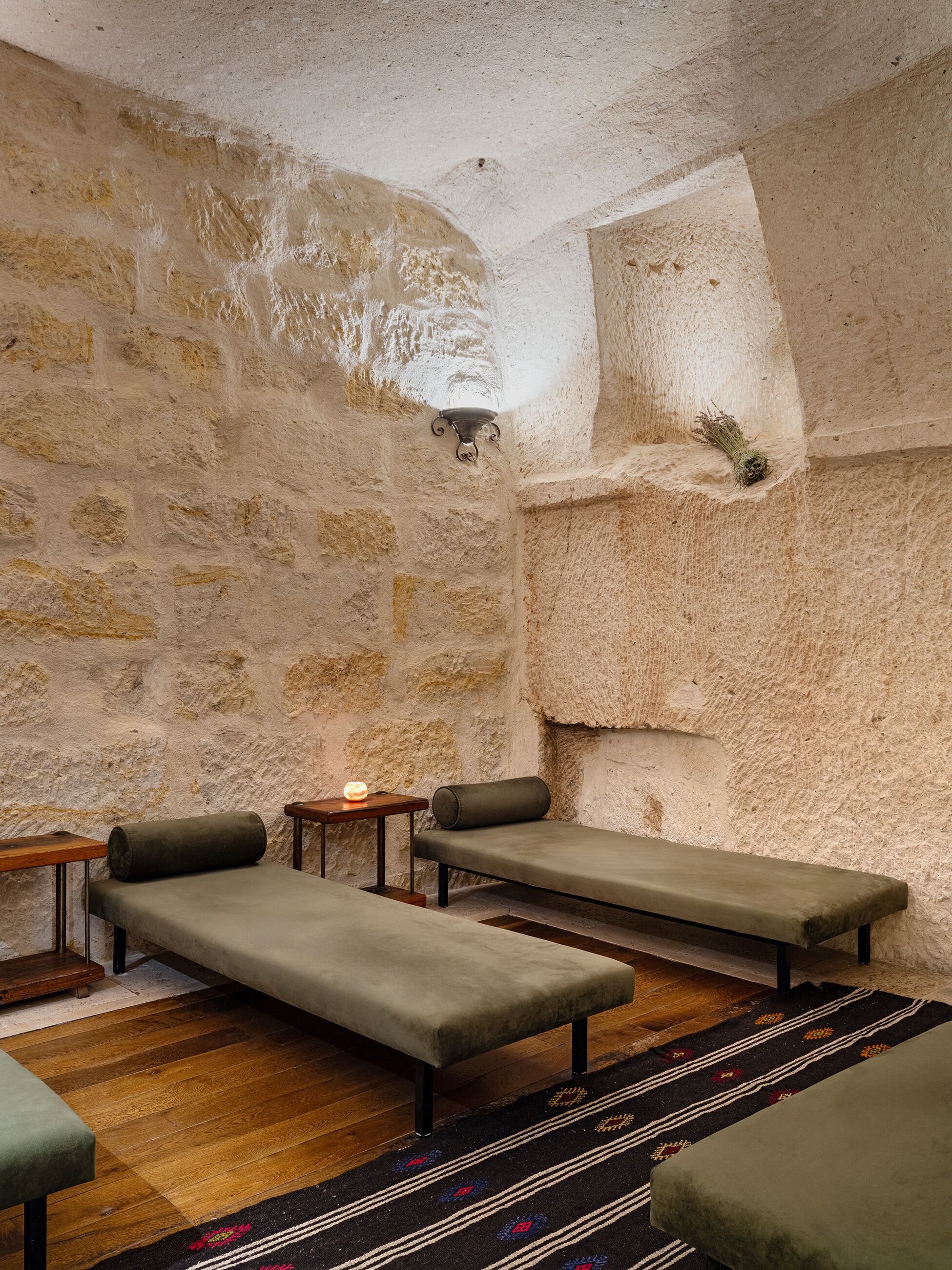 A stone-walled room with three cushioned benches, a small table with a candle, and a patterned rug.