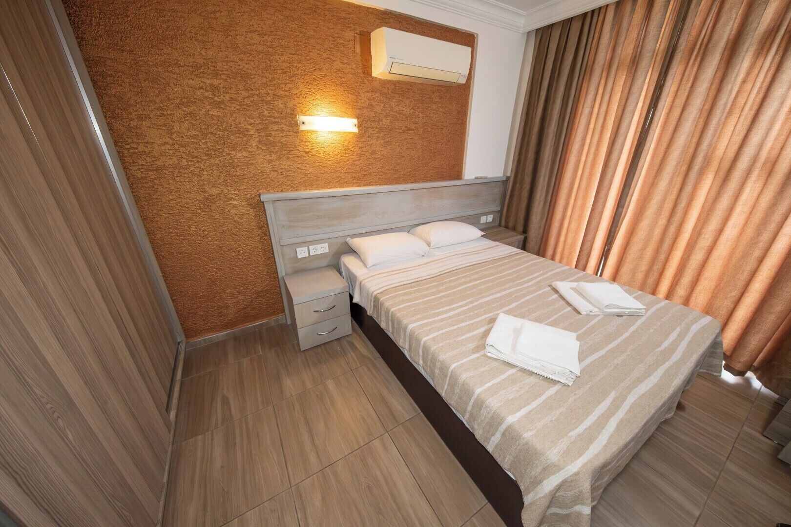 A hotel room with a bed, a nightstand, a wall-mounted air conditioner, and a window with curtains.