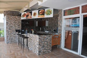 A kitchen with a central island, bar stools, a stone backsplash, and a dining area with a glass door. - Marina Apart Otel (Marmaris)