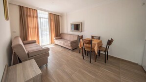 Una sala de estar con sofá, una mesa de comedor con sillas, un televisor montado en la pared y ventanas grandes con cortinas.