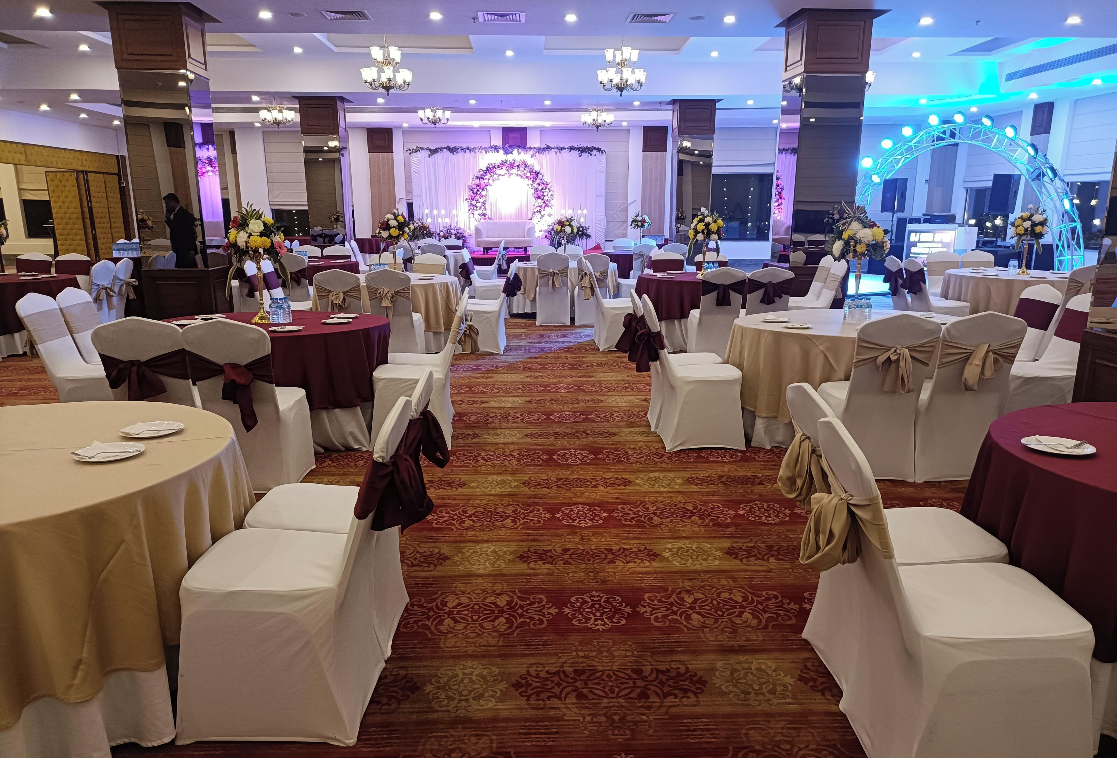 A banquet hall with round tables, white and burgundy chairs, and a stage with floral decorations.