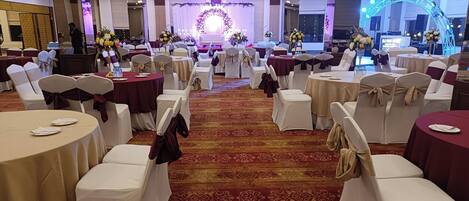 A banquet hall with round tables, white and burgundy chairs, and a stage with floral decorations.