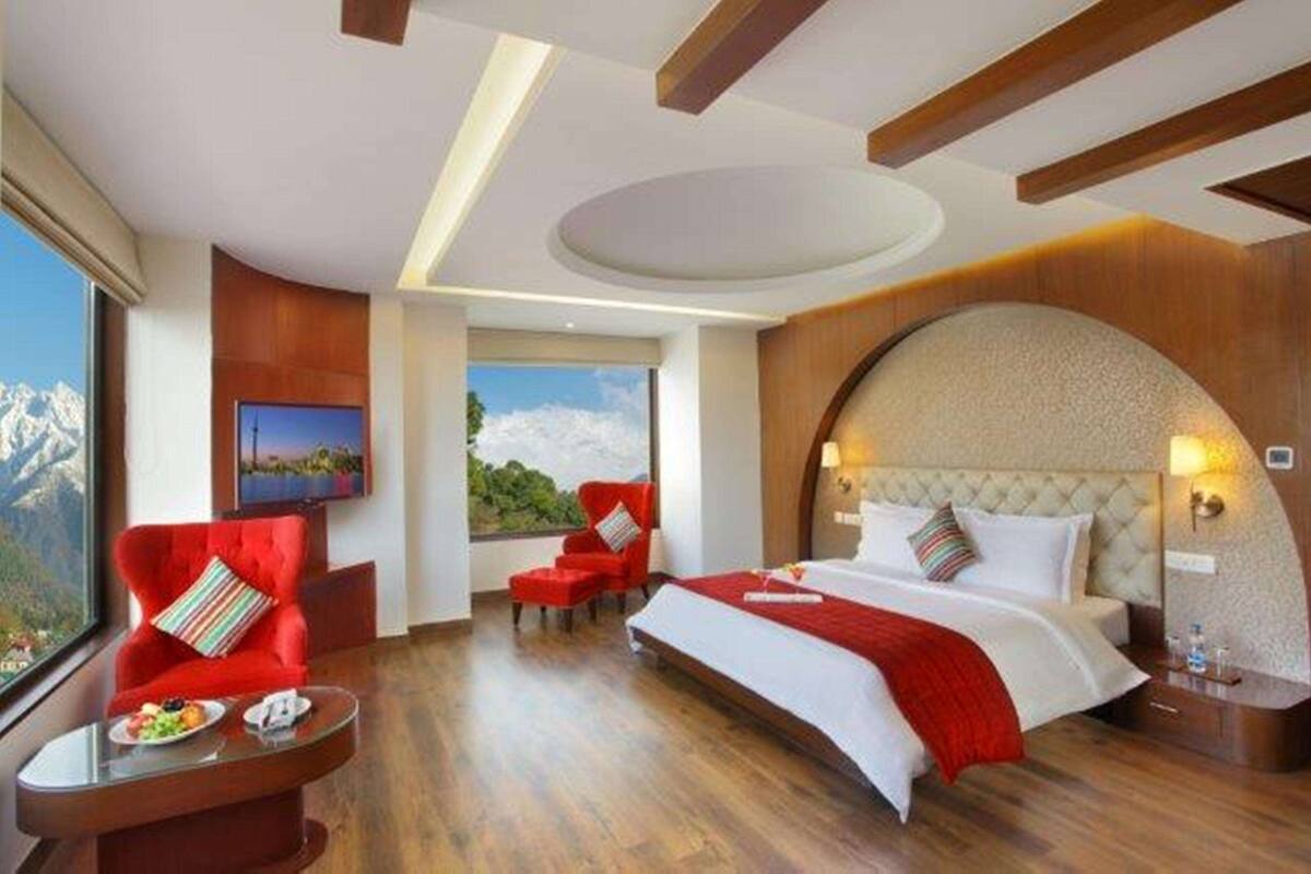 Ein Hotelzimmer mit einem großen Bett, einem roten Sessel, einem kleinen Tisch mit einer Obstplatte und Blick auf Berge durch das Fenster.