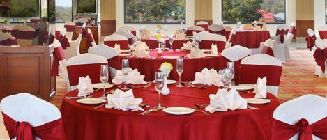 Un salón de banquetes con mesas redondas dispuestas para cenar, sillas blancas con cintas rojas y grandes ventanales con vistas al exterior.