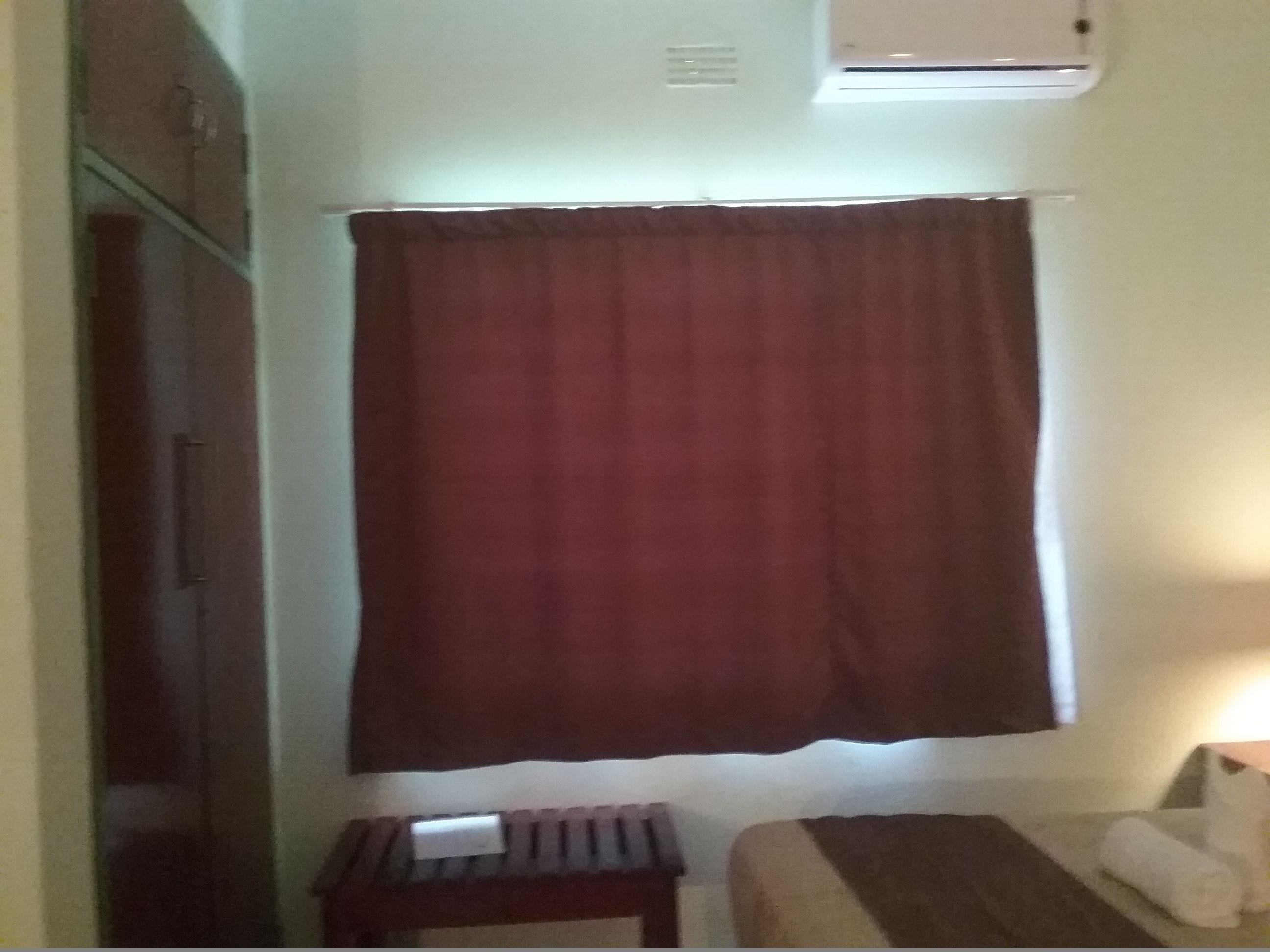 A room with a bed, a small table, a window with a curtain, and an air conditioning unit on the wall.