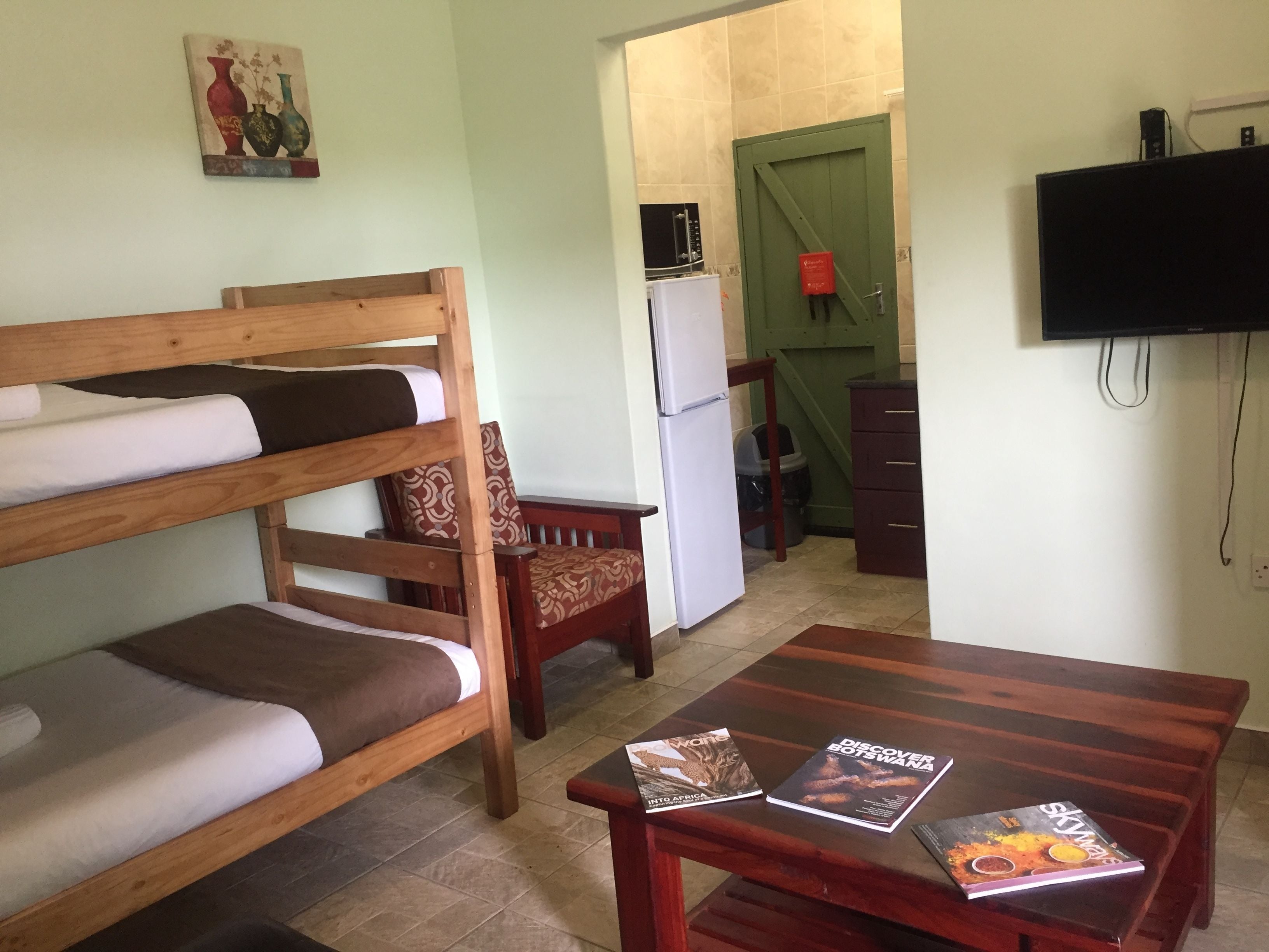 A room with bunk beds, a small wooden table with travel books, a television, and a kitchen area with a refrigerator and a green door.