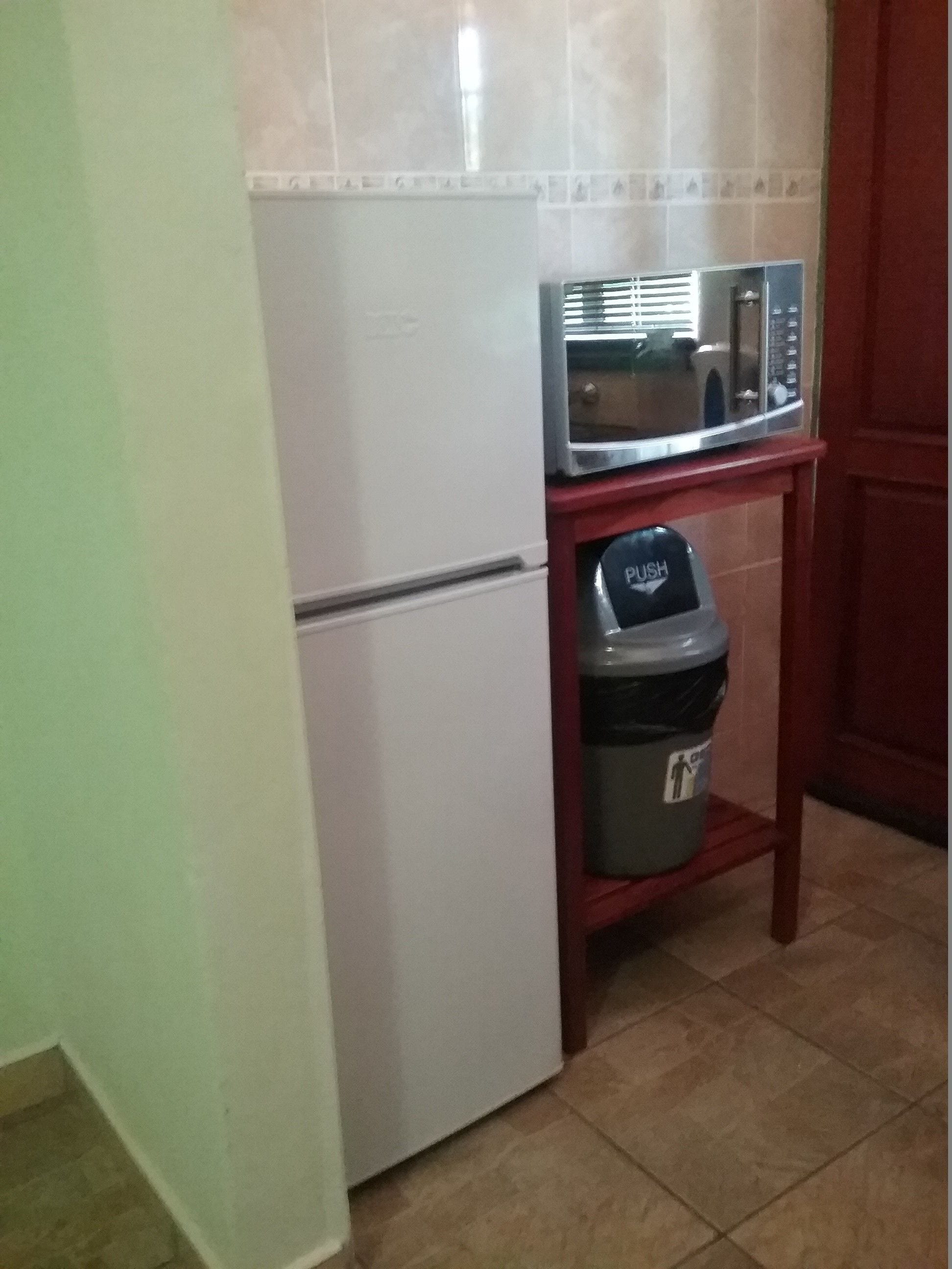 A compact kitchen with a white refrigerator, a microwave on a small wooden shelf, and a trash bin labeled "PUS."