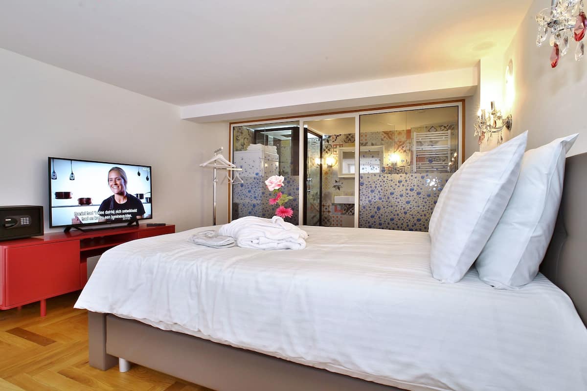 Quarto de hotel com cama, televisão, penteadeira vermelha e banheiro visível através de uma porta de vidro.