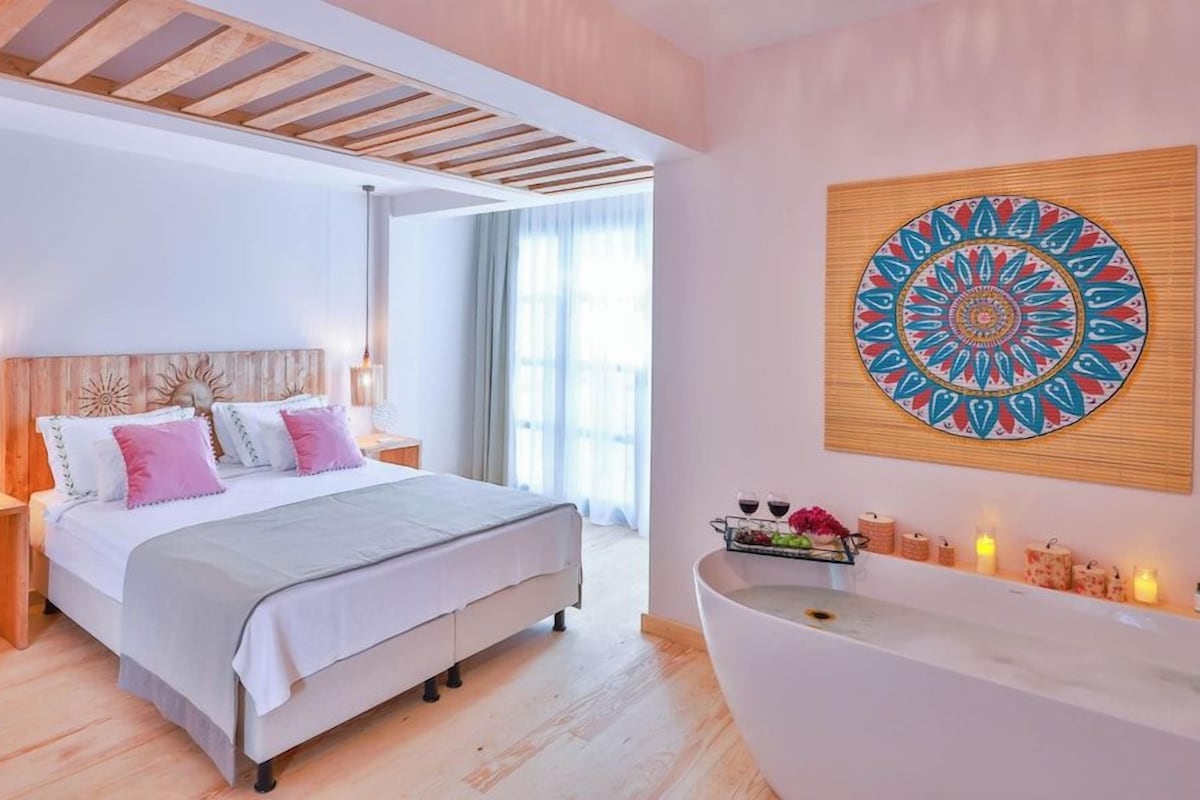 Ein Schlafzimmer mit einem Bett, Nachttischen, einem an der Wand befestigten Kunstwerk, einer Badewanne und einem Fenster mit Vorhängen.