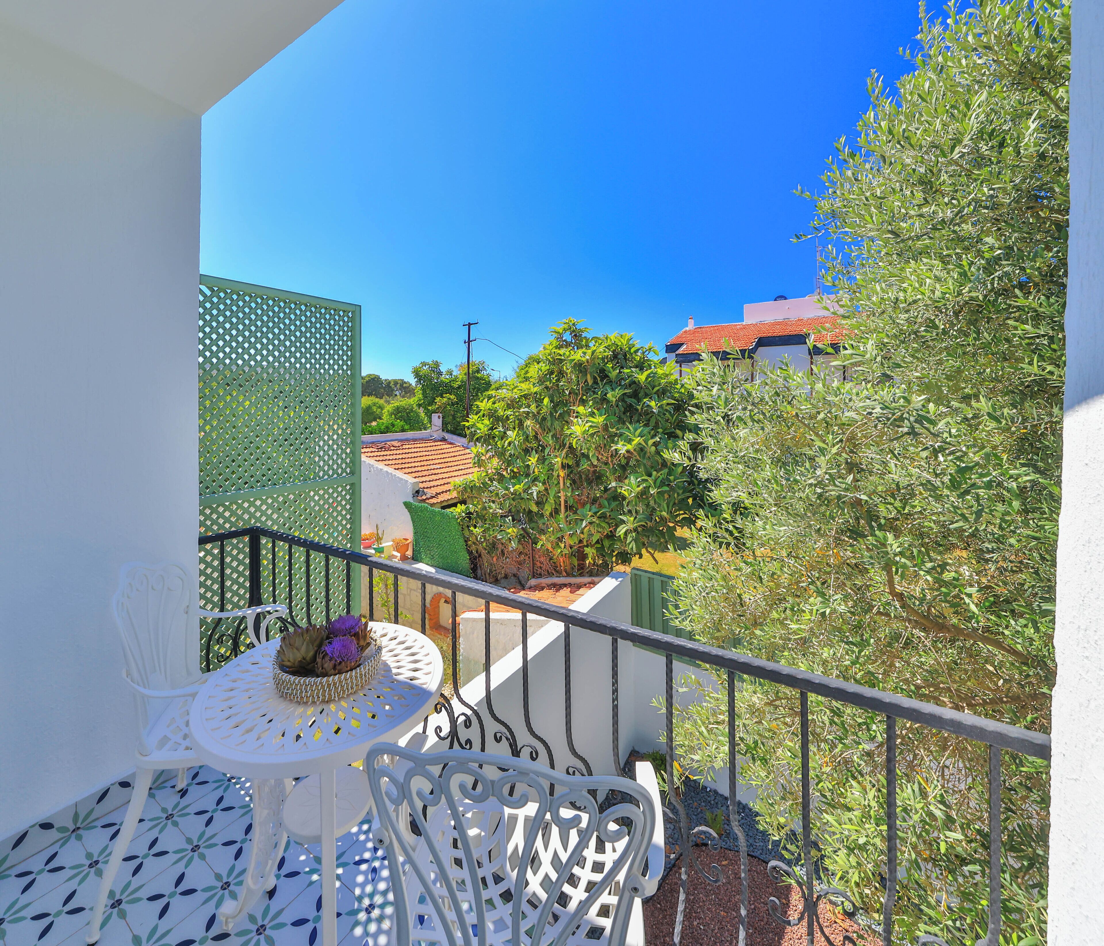 Un balcon avec une table et des chaises blanches, une clôture verte et une vue sur des immeubles et des arbres.