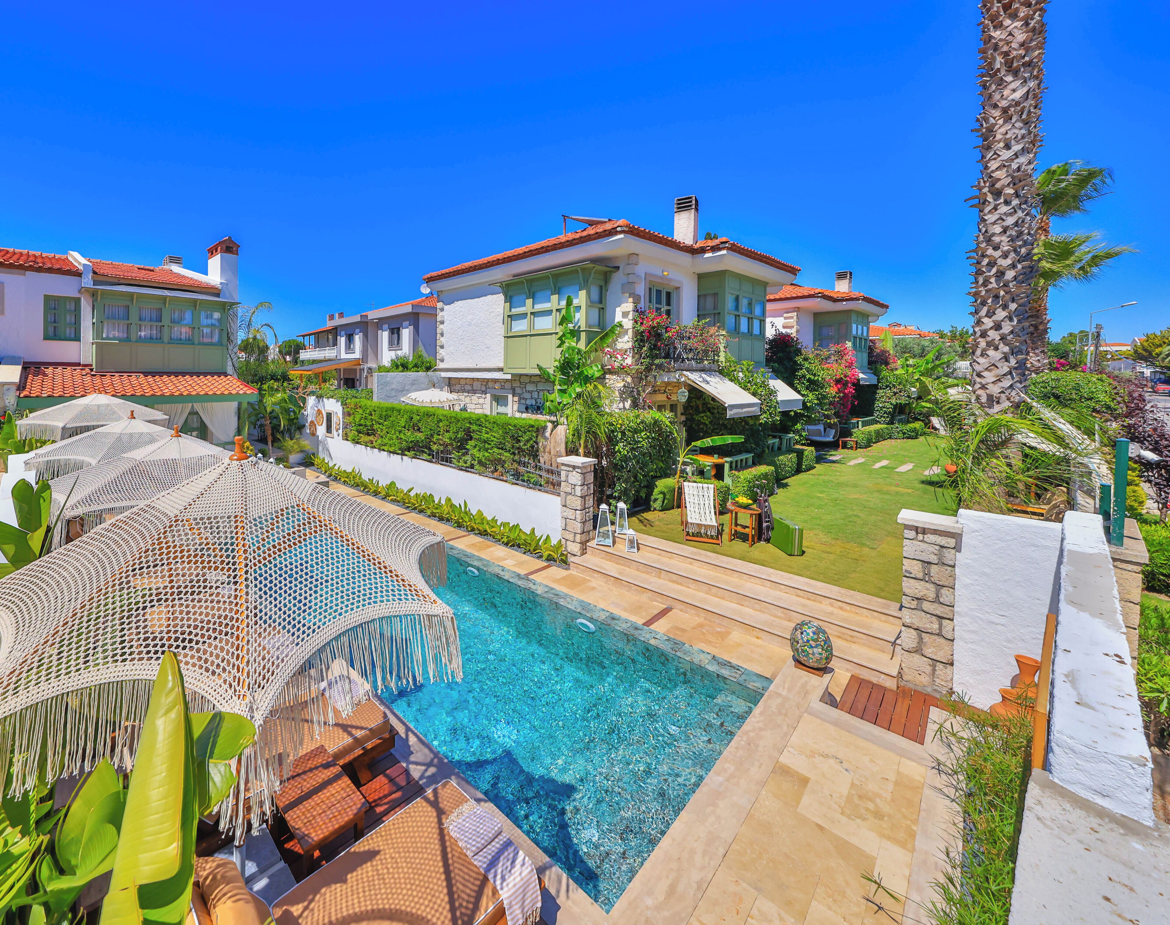 Un área junto a la piscina con sillones de descanso y sombrillas, una piscina azul cristalina y una casa grande con techo de tejas rojas.