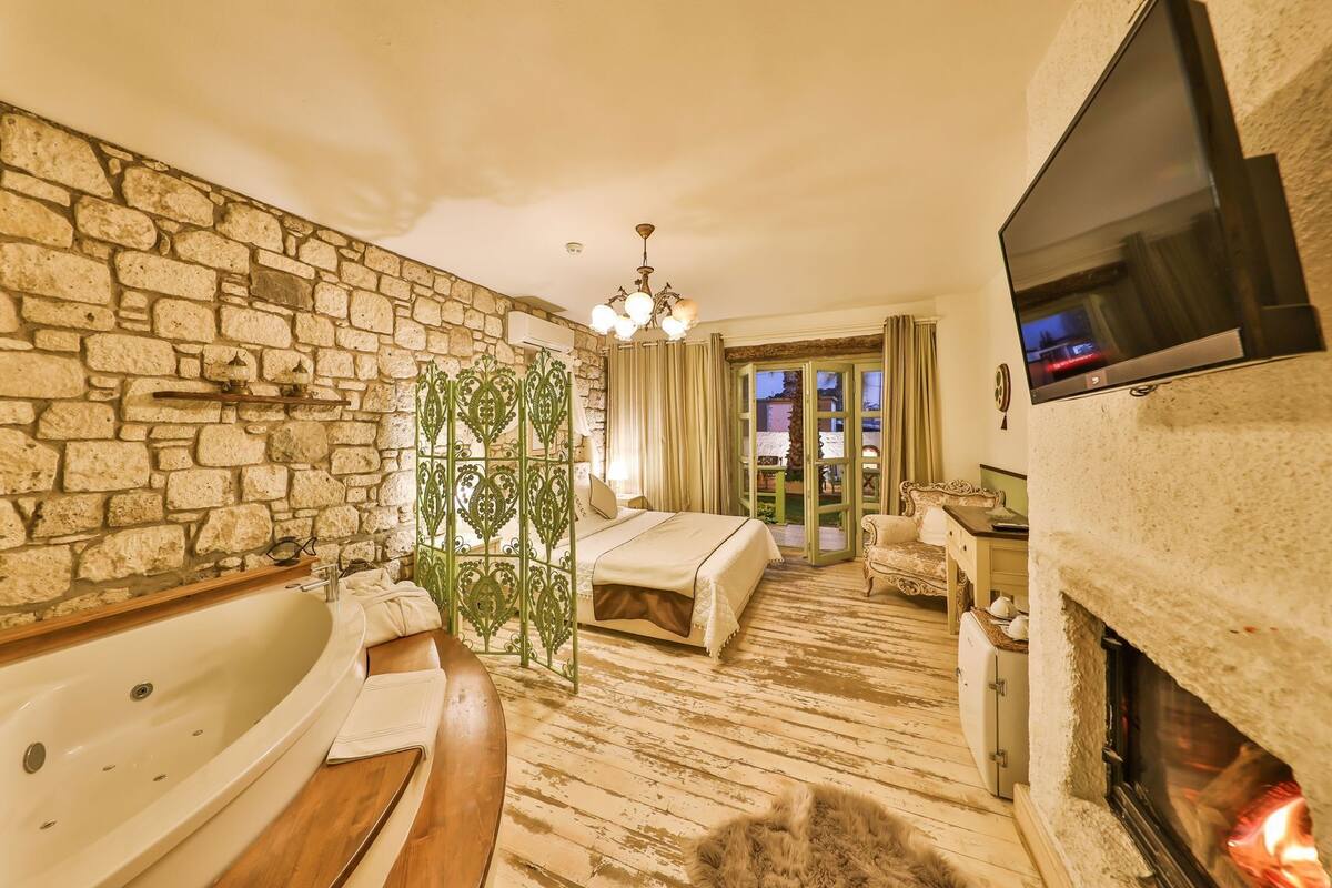 A hotel room with a stone wall, a large bed, a bathtub, a fireplace, and a television.