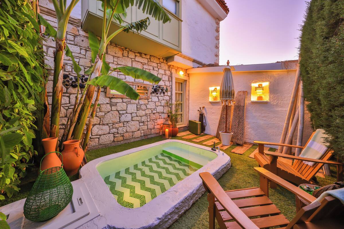 A stone building with a small pool, wooden chairs, and potted plants.