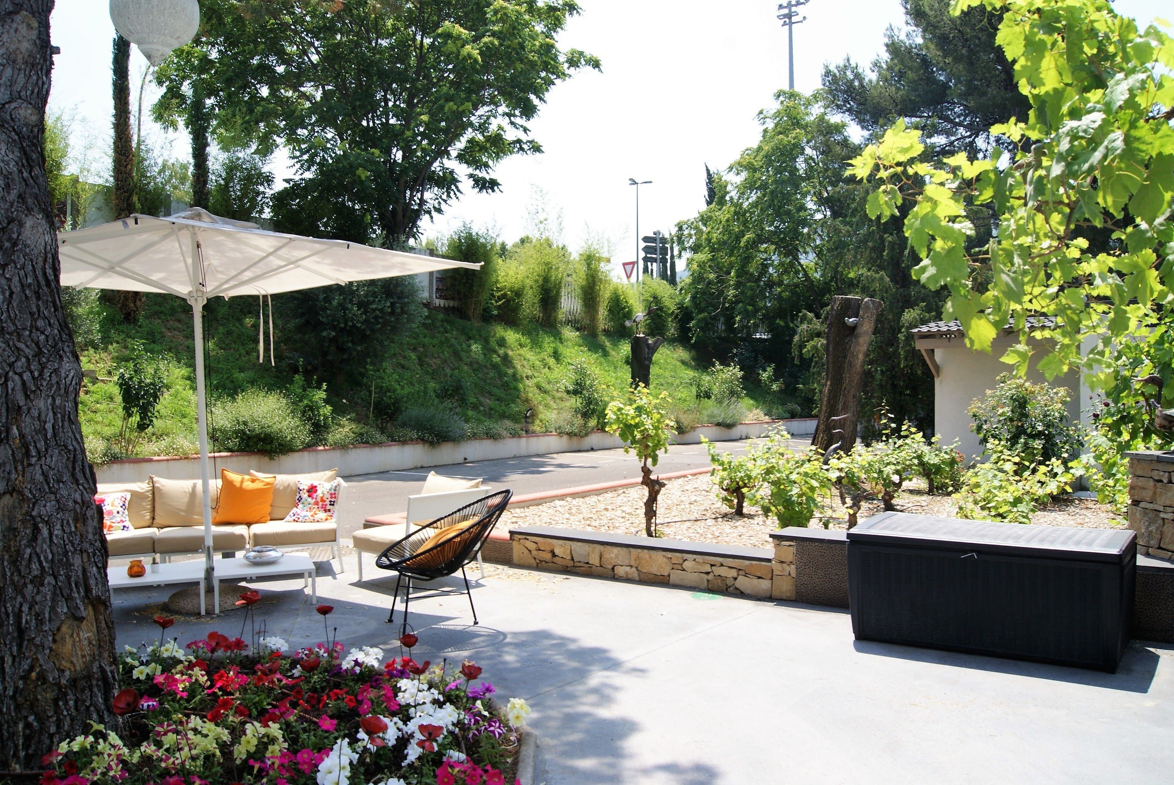 An outdoor patio with a white umbrella, a sofa with orange cushions, a black chair, and a planter with flowers.
