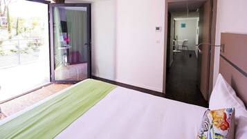 Habitación de hotel con cama, alfombra verde, almohada colorida, puerta corrediza de vidrio que da a un balcón y un pasillo con una silla y una mesa.