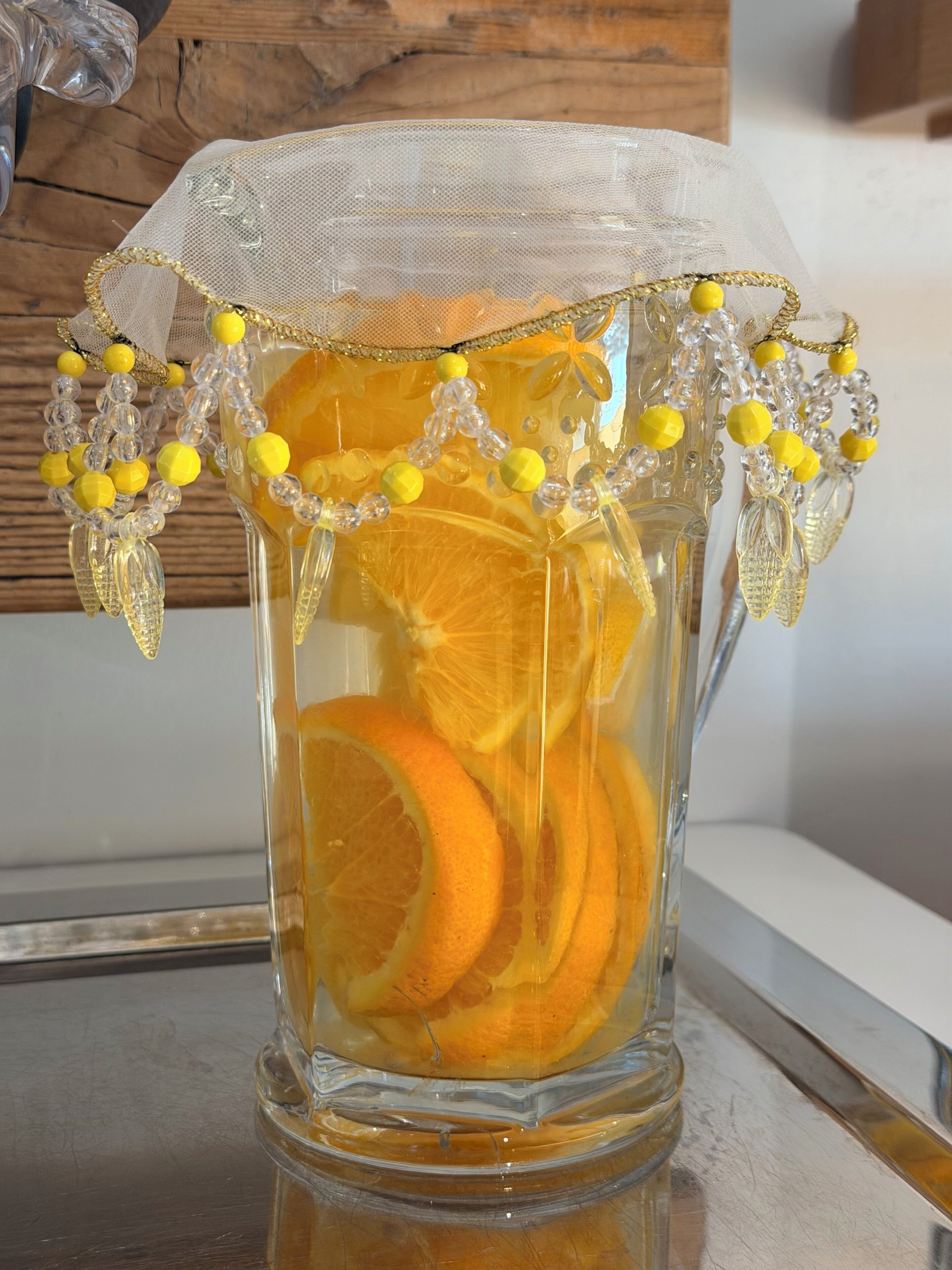 A clear glass pitcher with a decorative lace cover and yellow beads, containing sliced oranges.
