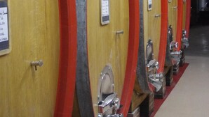 Una fila de barricas de madera con aros metálicos y grifos de metal, probablemente usadas para añejar vino.