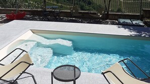 Zona de piscina con sillones reclinables y una mesa, con vistas a un viñedo y edificios a lo lejos.