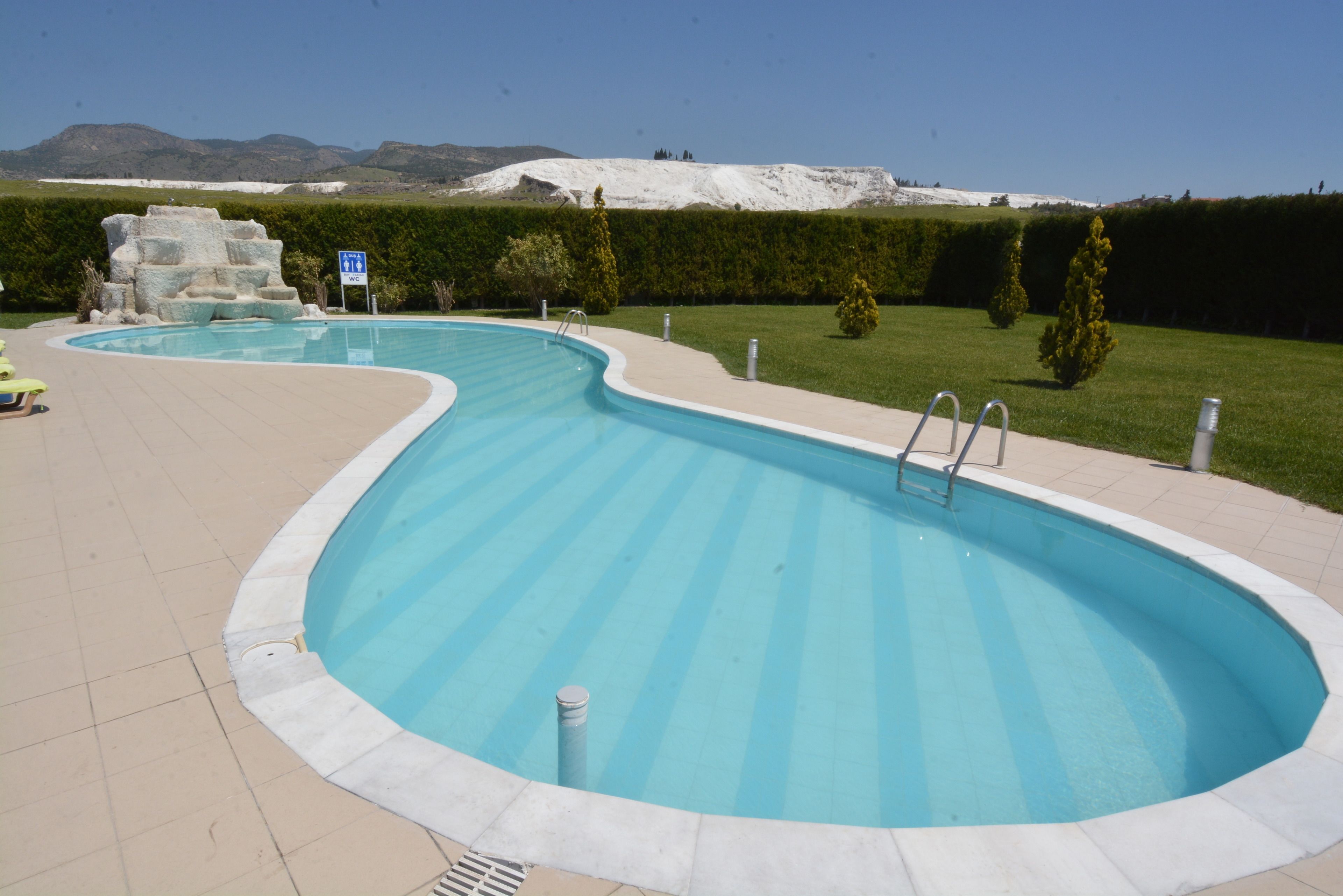 Una piscina con una estructura de piedra, cielo despejado y una cadena montañosa distante.