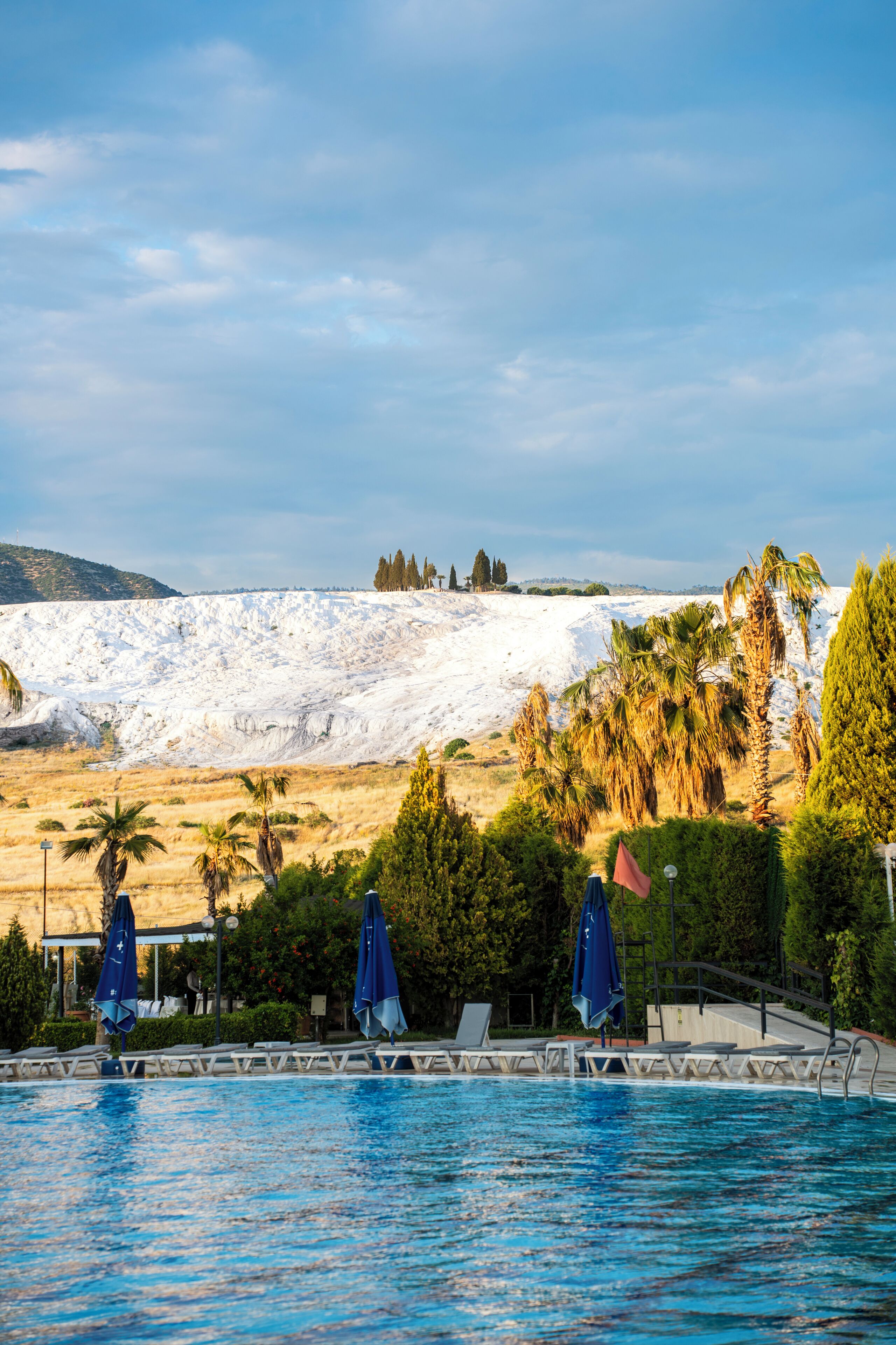 Une piscine avec des chaises longues et des parasols, une colline enneigée en arrière-plan et un ciel dégagé.