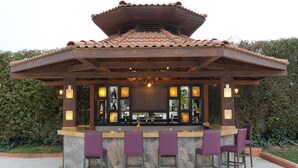Uma área de bar ao ar livre coberta, com telhado de azulejos, banquetas e um balcão cheio de garrafas.