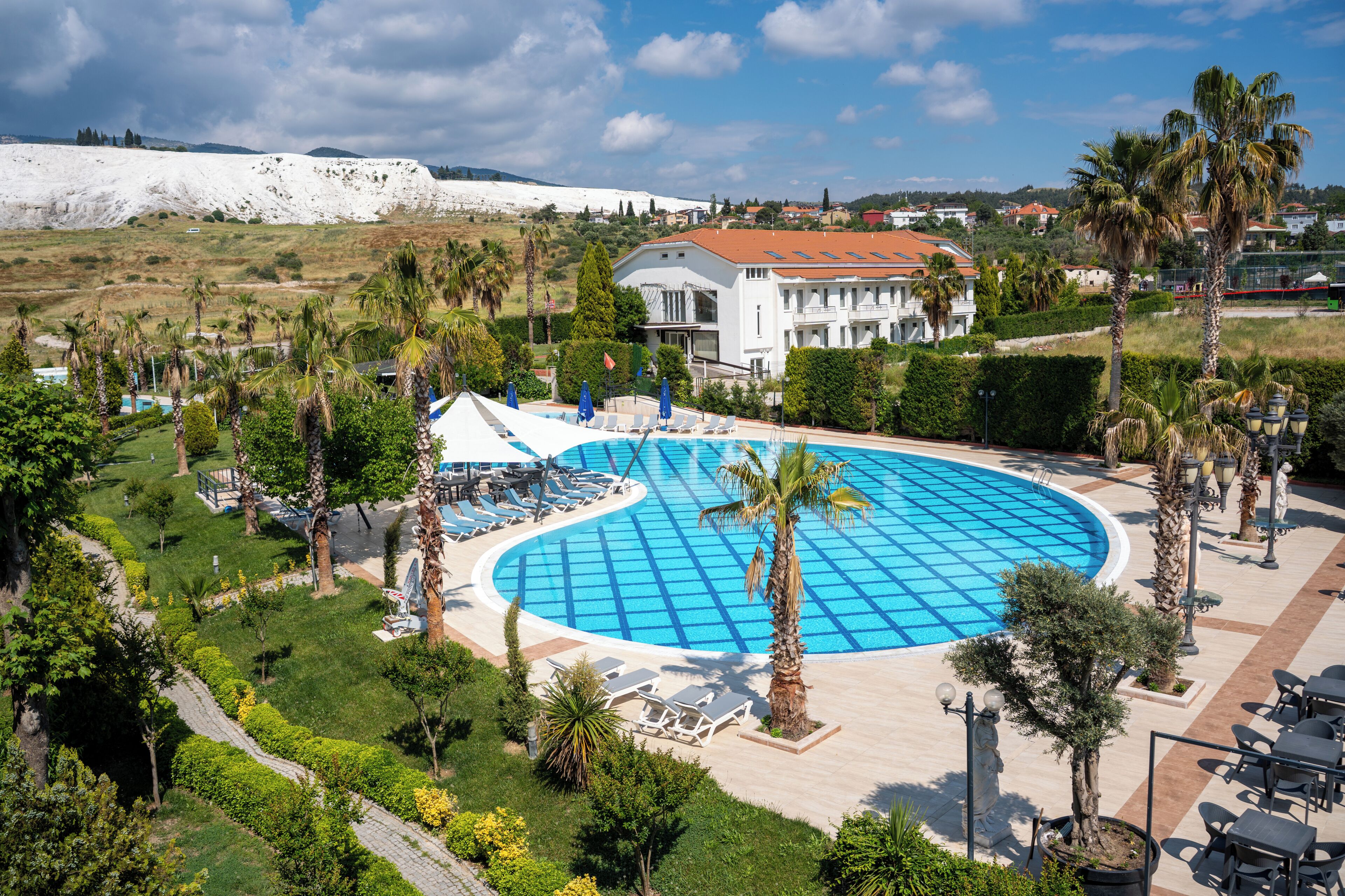 A large swimming pool surrounded by palm trees and lounge chairs, with a white building in the background.