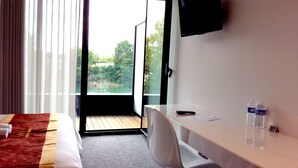 A hotel room with a bed, a desk, a chair, a TV, and a view of a garden through a large window. - Hôtel de la seine (Epinay-sur-Seine)