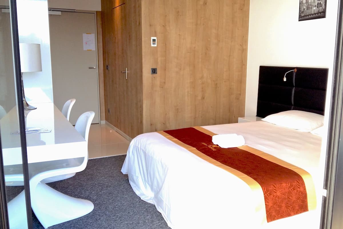 A hotel room with a bed, a desk with chairs, a wooden wardrobe, and a wall-mounted TV.