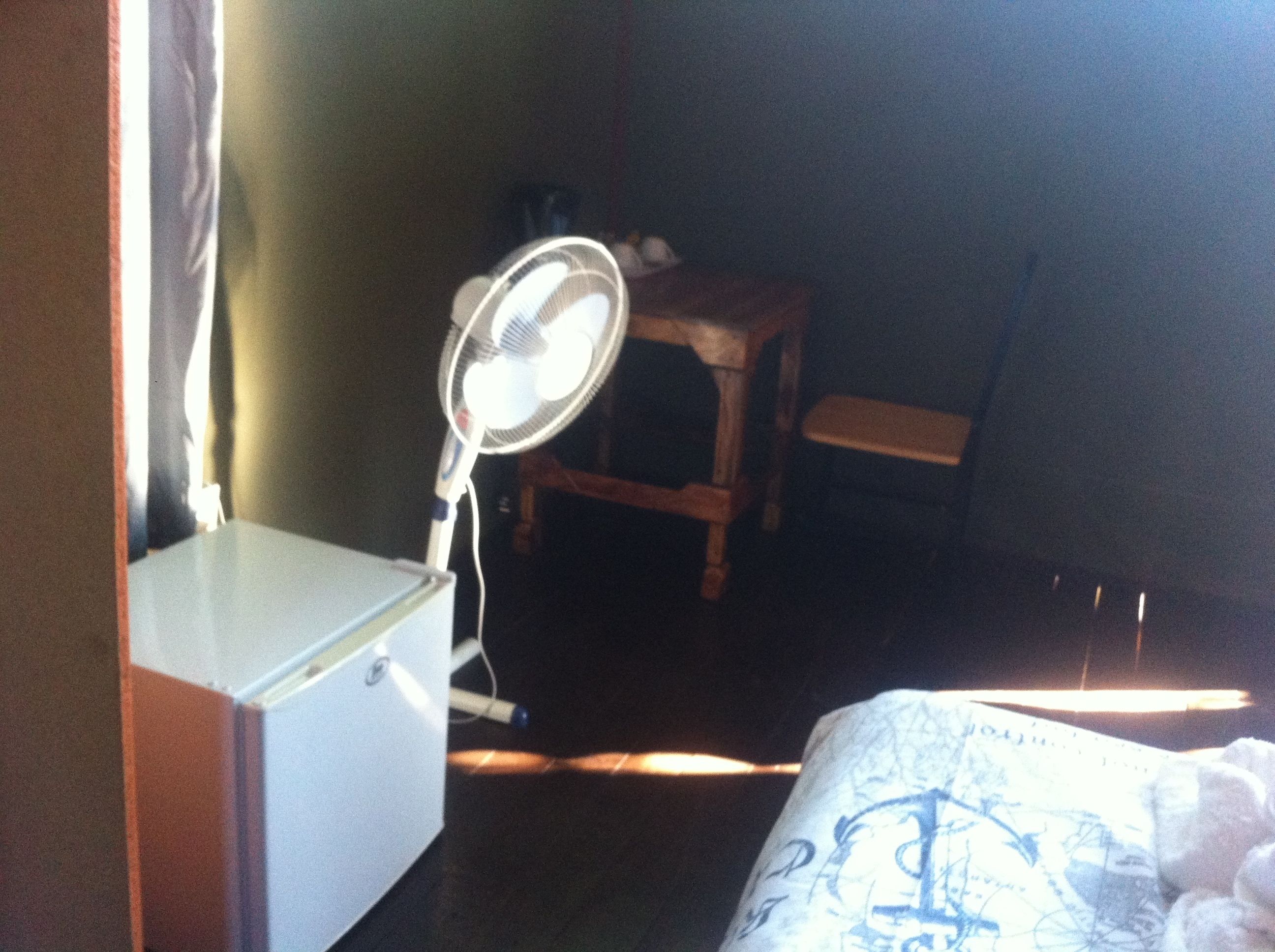 Una stanza con un letto, un comodino, una sedia, una scrivania e una ventola da terra.