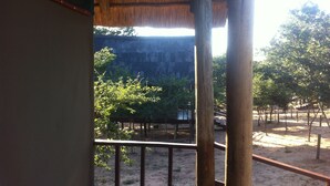 Vista desde un balcón con techo de paja, sobre una zona arenosa con árboles y un acantilado rocoso.